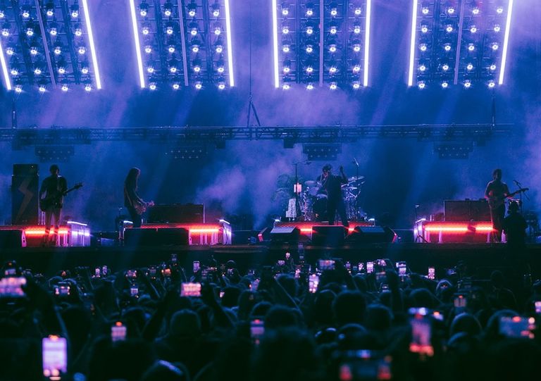 Rock band The Strokes performing live on a concert stage under dramatic blue and purple lights in front of a cheering crowd holding up mobile phones.